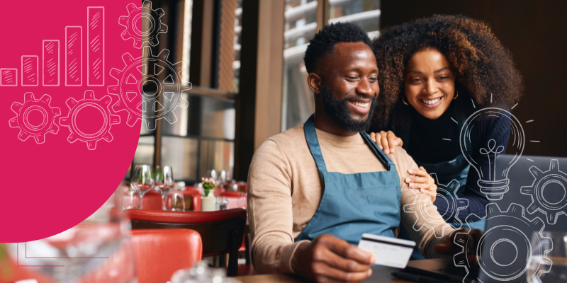 Um homem negro de avental azul e camiseta de manga longa beje e uma mulher negra com uma blusa azul segurando um catão branco sentado em uma mesa se um restaurante em frente a um notebook.