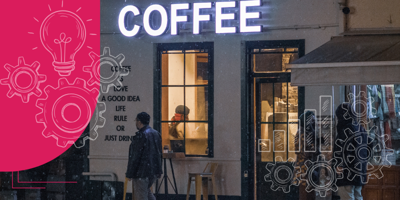 Café moderno com janela de vidro, clientes nas mesas, baristas, plantas, iluminação quente e mesas de madeira.