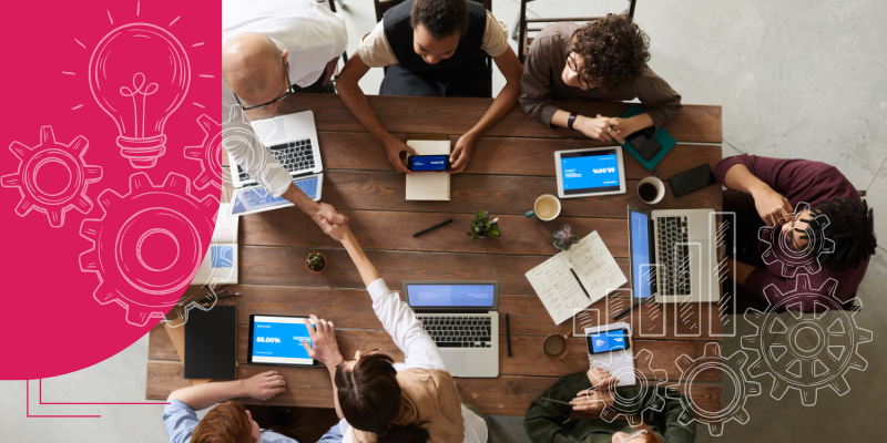 Pessoas diversas em torno de uma mesa de madeira com laptops e plantas ao redor em um escritório moderno.